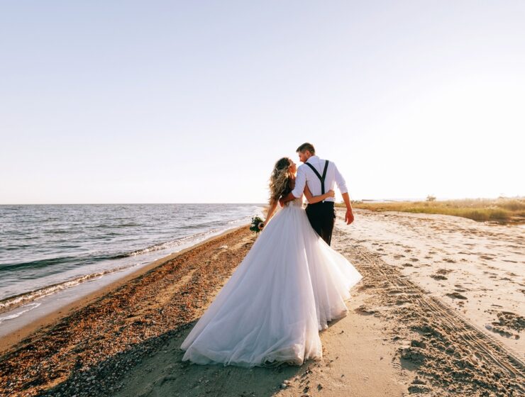 A bride and groom walking along a sandy shore with their arms around one another’s waist on a sunny day.