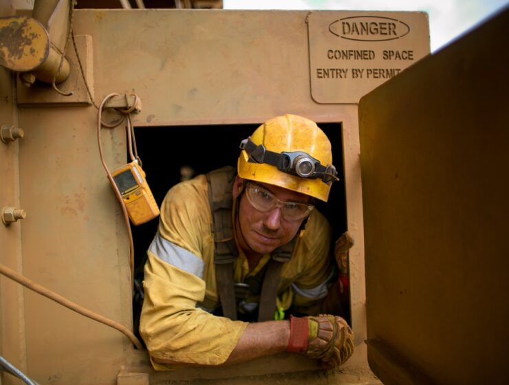 A maintenance worker wearing reflective PPE and a helmet comes out of a confined space where he was working.