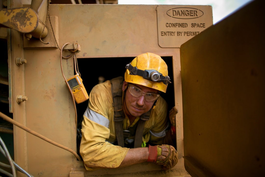 A maintenance worker wearing reflective PPE and a helmet comes out of a confined space where he was working.