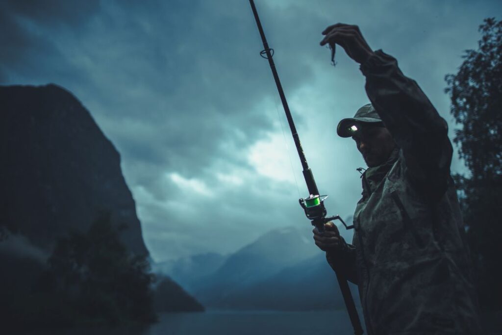 A man in fishing gear and a headlamp stands with a fishing rod. It's nighttime, and he holds the hook in his other hand.