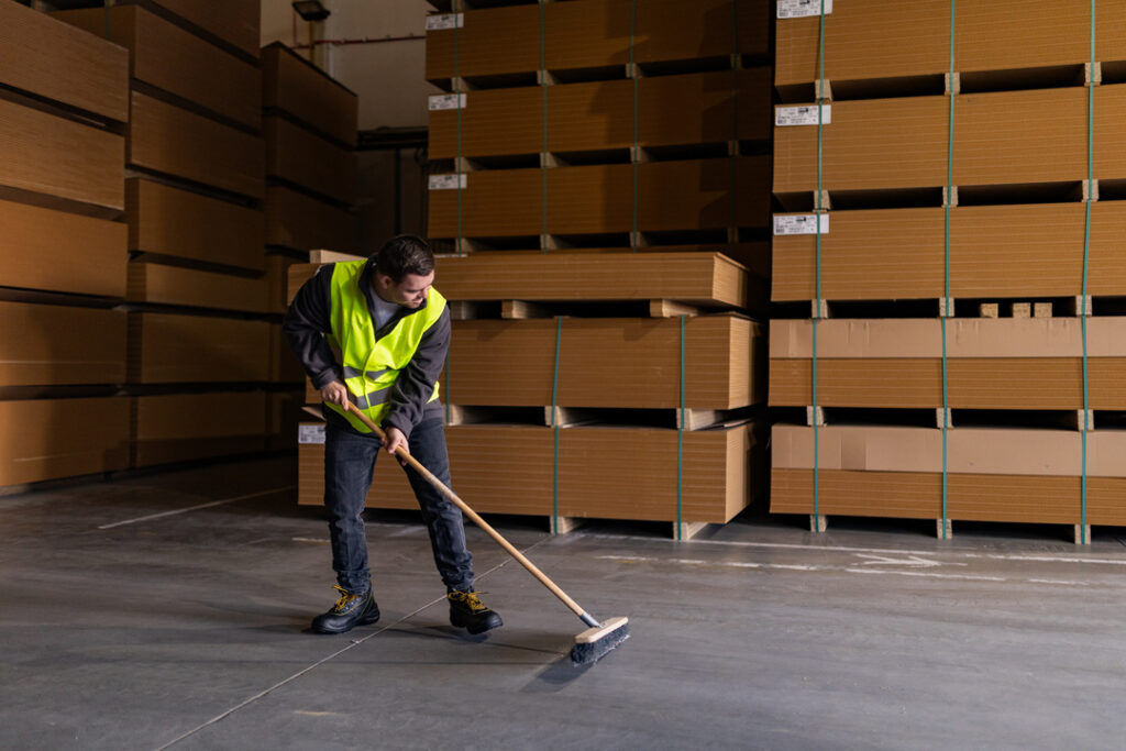 A man wearing a yellow safety vest sweeps the floor of a warehouse. Palettes of materials are stacked behind him.