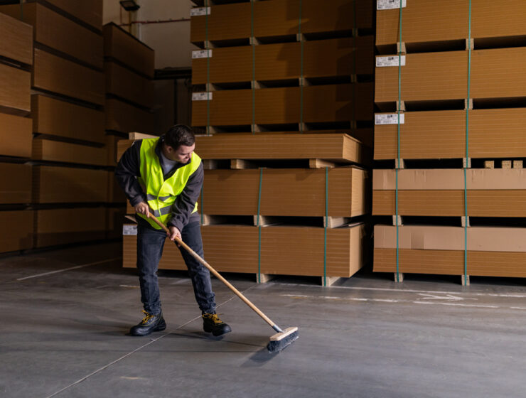 A man wearing a yellow safety vest sweeps the floor of a warehouse. Palettes of materials are stacked behind him.