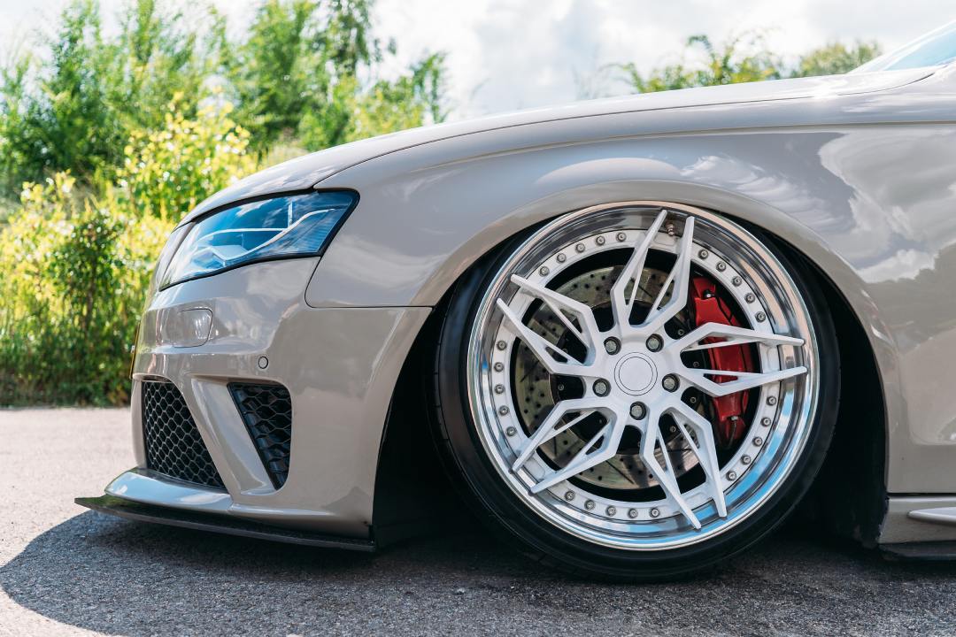 A lowered beige car with polished alloy wheel, red brake caliper, and sporty bumper parked on outdoor pavement.
