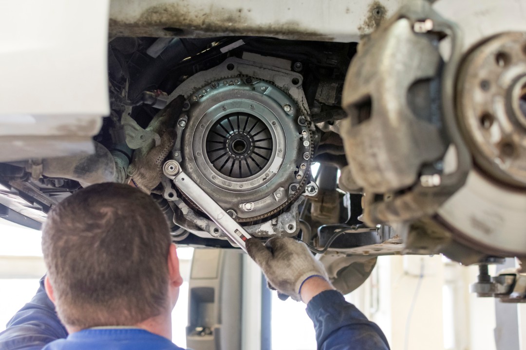 A rear view of a mechanic working on the clutch system of a vehicle elevated on a lift with a socket wrench.