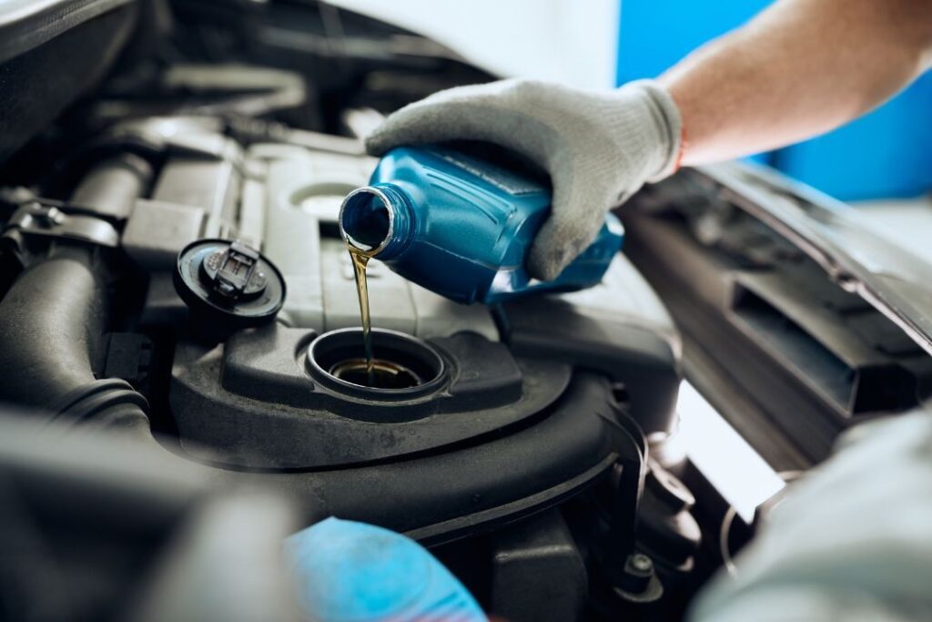 A close-up of a gloved hand holding a blue bottle and pouring an amber liquid into a reservoir in a car's engine.