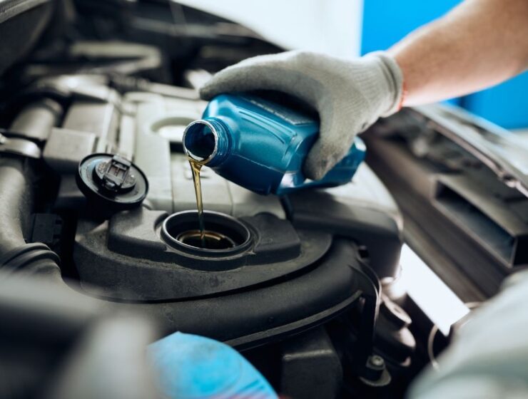 A close-up of a gloved hand holding a blue bottle and pouring an amber liquid into a reservoir in a car's engine.