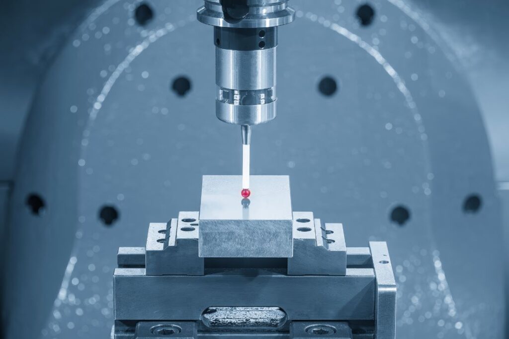 A stylus probe with a ruby tip measures a metal block clamped in a fixture inside a CMM chamber.
