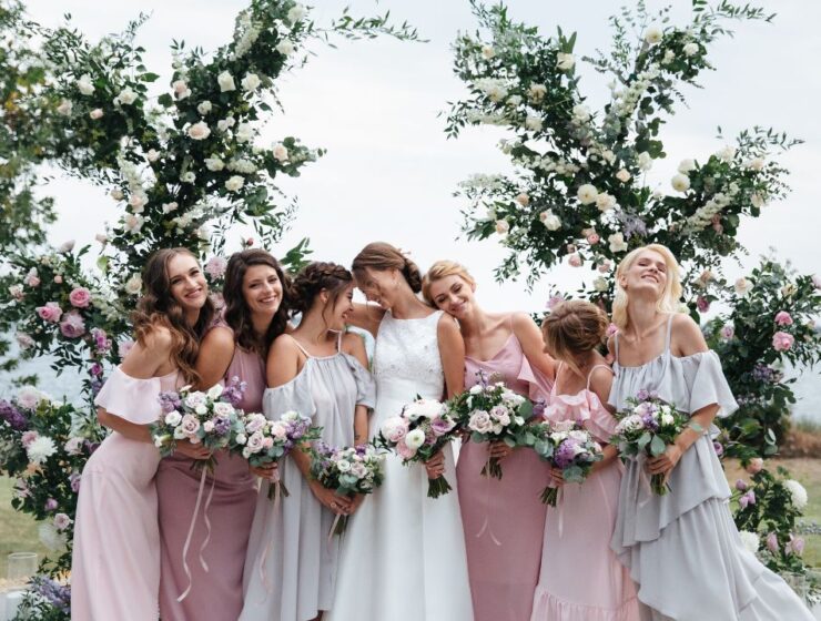 A group of smiling bridesmaids in soft pink and beige dresses holding bouquets, standing together under a floral arch.