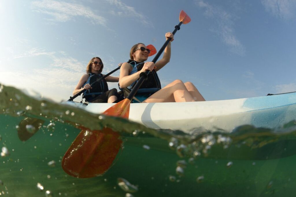 A low-angle view of two women in lifejackets kayaking on clear water. They paddle in unison beneath a bright blue sky.