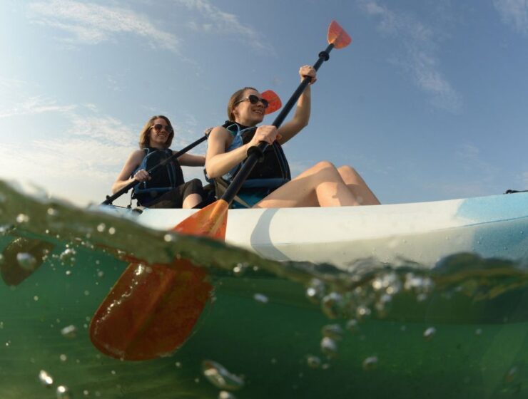 A low-angle view of two women in lifejackets kayaking on clear water. They paddle in unison beneath a bright blue sky.
