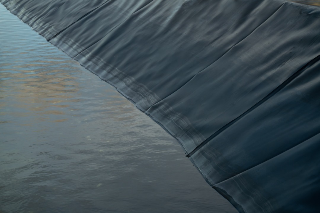 a black pond liner draping along the edge of murky water with folds and seams visible across the surface.
