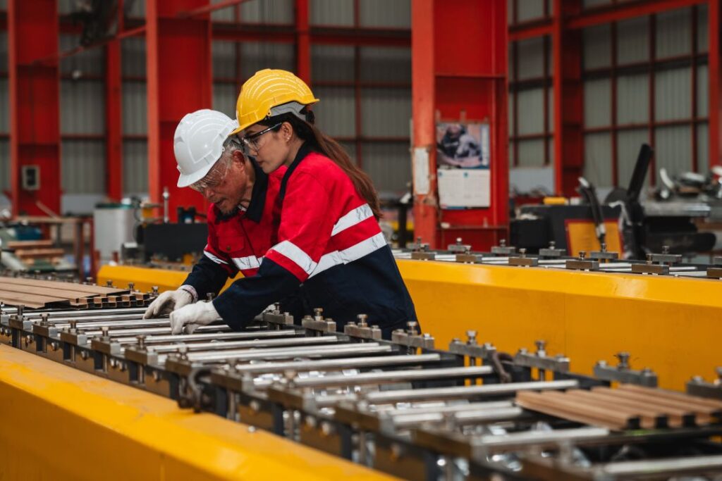 Two workers in hard hats and safety jackets stand in front of a roller conveyor. They inspect the rollers.