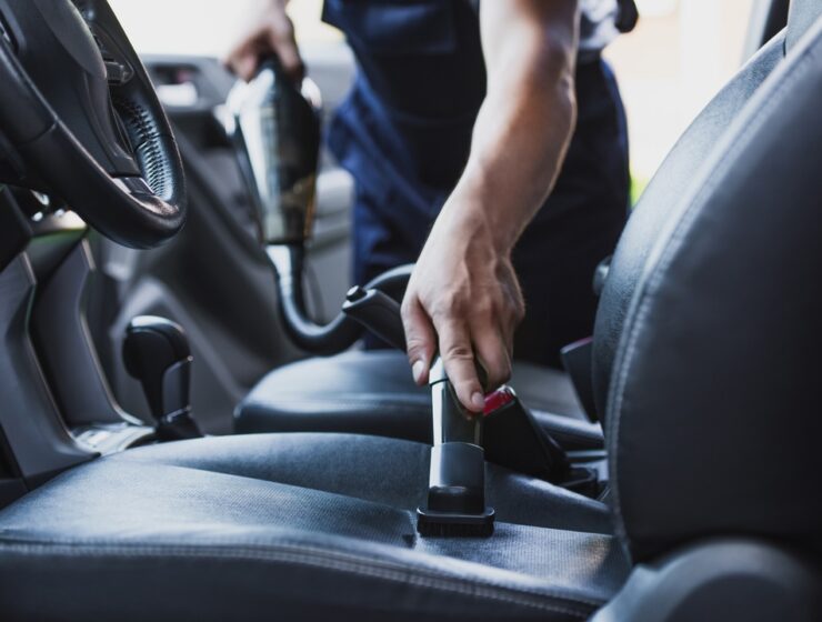 A person using a vacuum hose cleans a dark fabric driver's seat beside a steering wheel and center console.