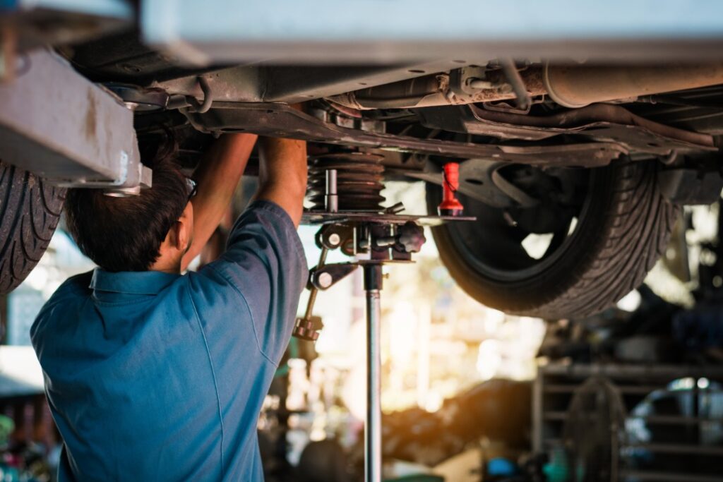 A person in a blue shirt working beneath a raised car near tires, suspension parts, and a red flashlight.