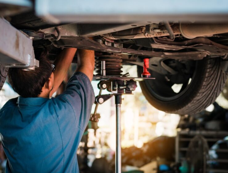 A person in a blue shirt working beneath a raised car near tires, suspension parts, and a red flashlight.