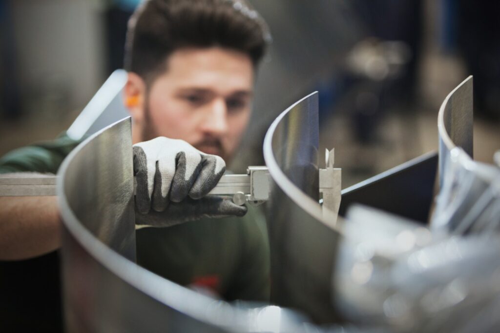 A close-up of a worker wearing gloves measuring curved steel pieces with calipers inside a metal fabrication shop.