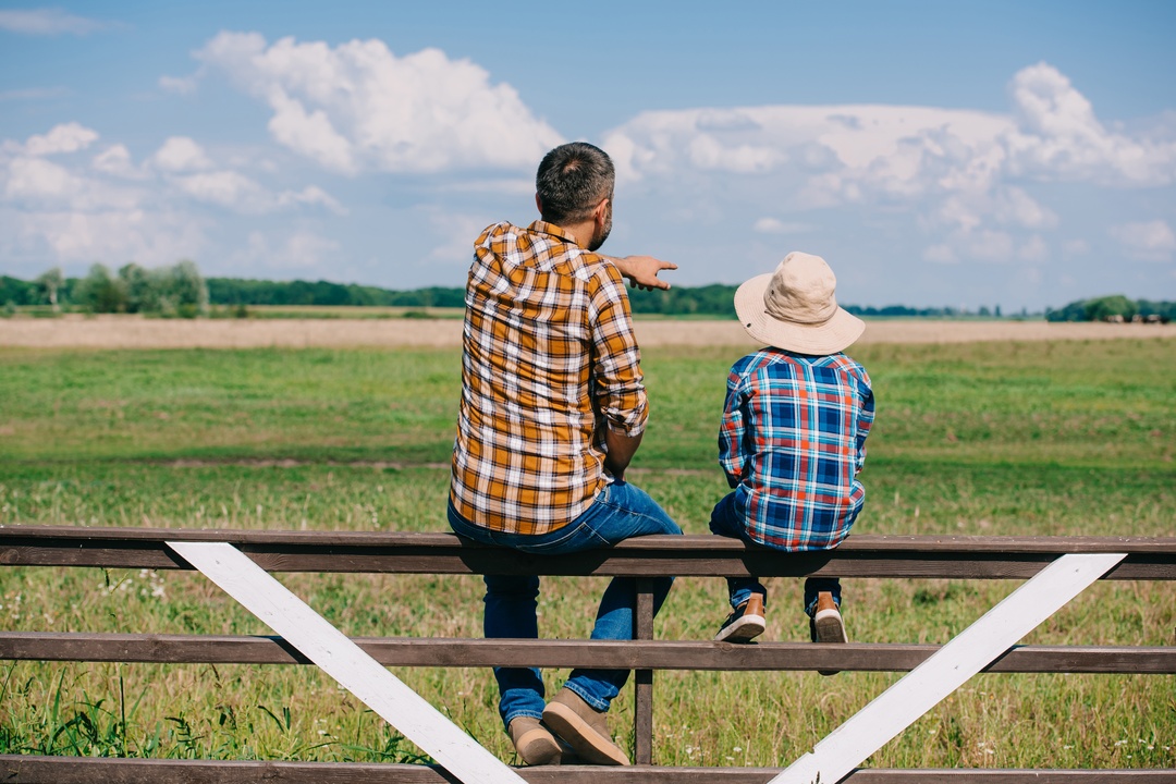 A father and son sitting on a wooden fence, facing a green field under a blue sky with large white clouds.
