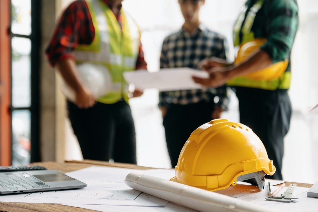 A close-up of a table with a hard hat, laptop, and construction blueprints on it while workers huddle in the background.
